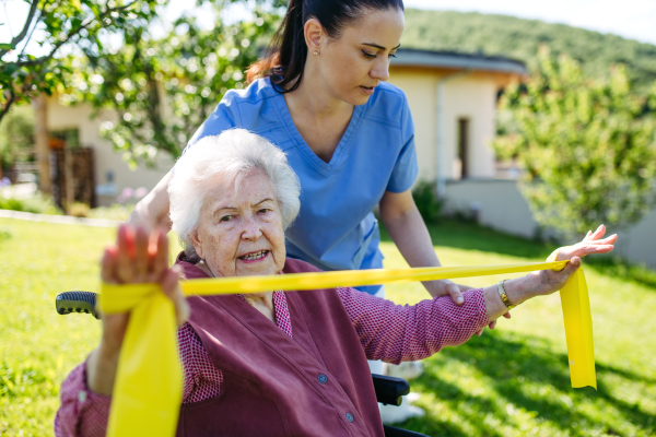 Female caregiver doing motorized exercises with senior woman in wheelchair. Nurse and elderly woman working out with yellow resistance band.