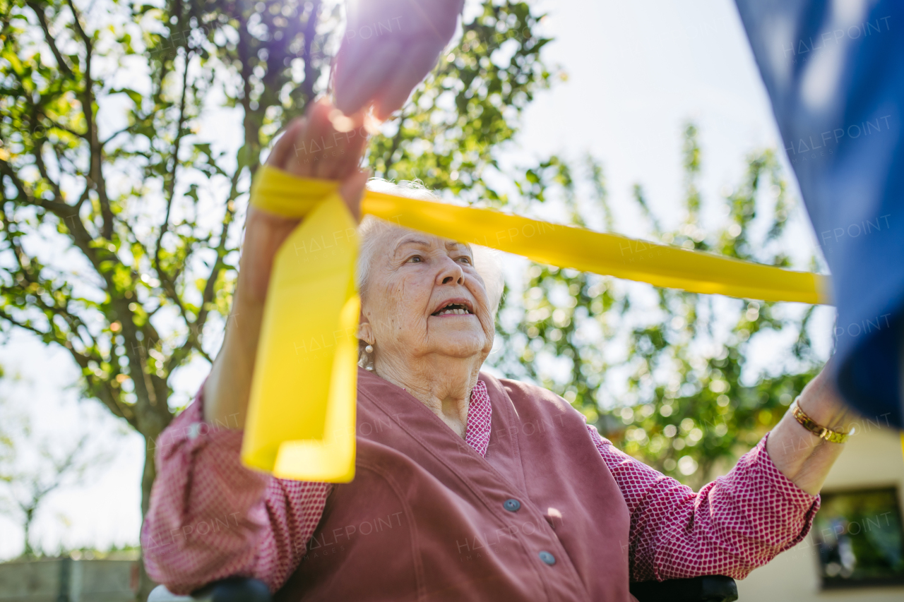 Female caregiver doing motorized exercises with senior woman in wheelchair. Nurse and elderly woman working out with yellow resistance band.