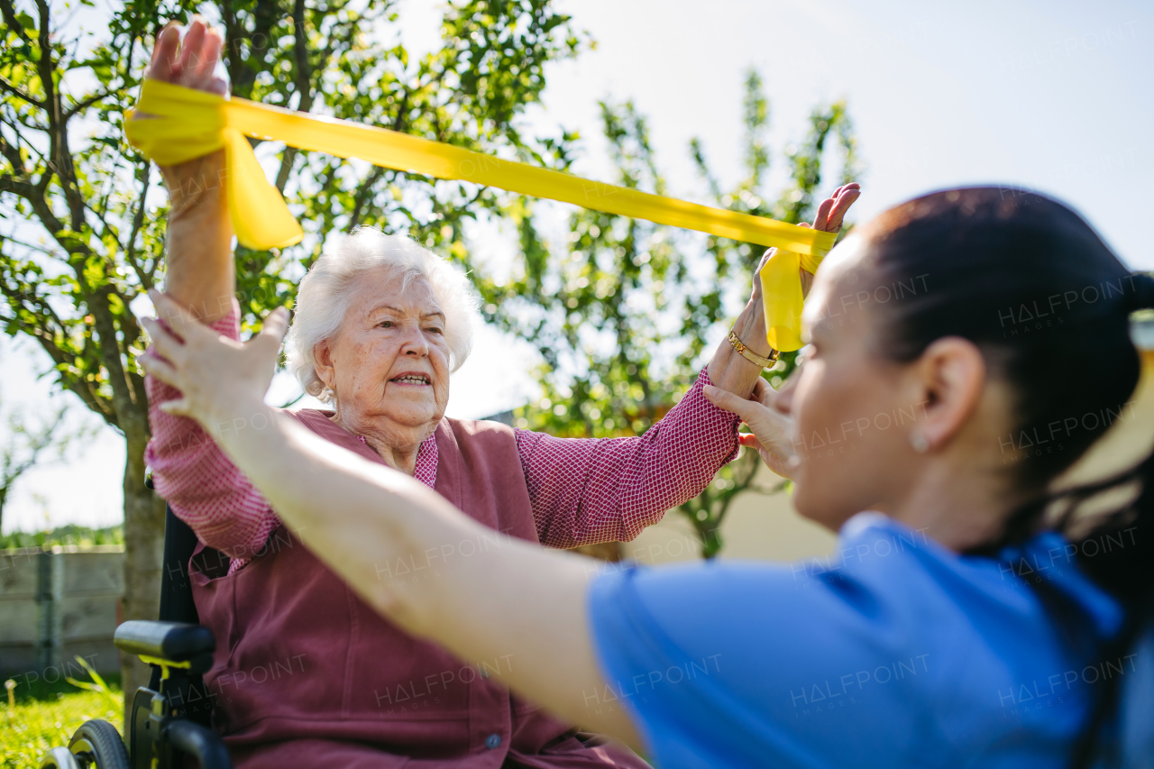 Female caregiver doing motorized exercises with senior woman in wheelchair. Nurse and elderly woman working out with yellow resistance band.