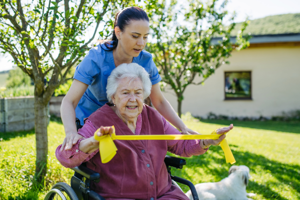 Female caregiver doing motorized exercises with senior woman in wheelchair. Nurse and elderly woman working out with yellow resistance band.