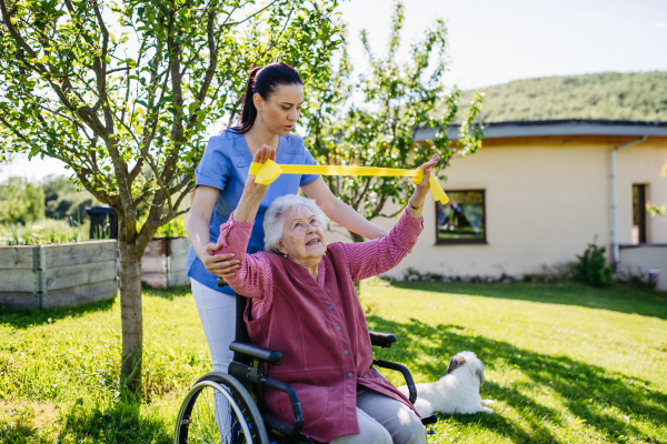 Female caregiver doing motorized exercises with senior woman in wheelchair. Nurse and elderly woman working out with yellow resistance band.