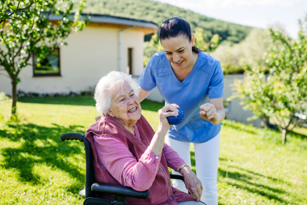 Female caregiver doing motorized exercises with senior woman in wheelchair. Nurse and elderly woman squeezing small soft ball.