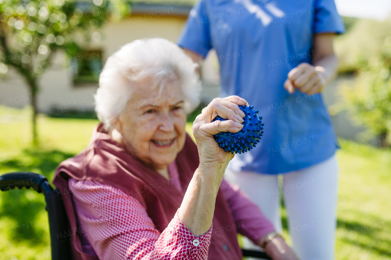 Female caregiver doing motorized exercises with senior woman in wheelchair. Nurse and elderly woman squeezing small soft ball.