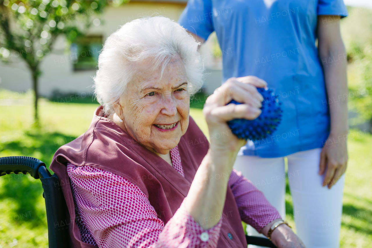 Female caregiver doing motorized exercises with senior woman in wheelchair. Nurse and elderly woman squeezing small soft ball.