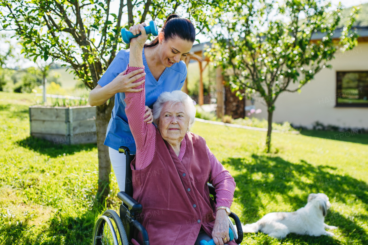 Female caregiver doing motorized exercises with senior woman in a wheelchair. Elderly woman working out with small dumbbell, nurse leading and teaching her.