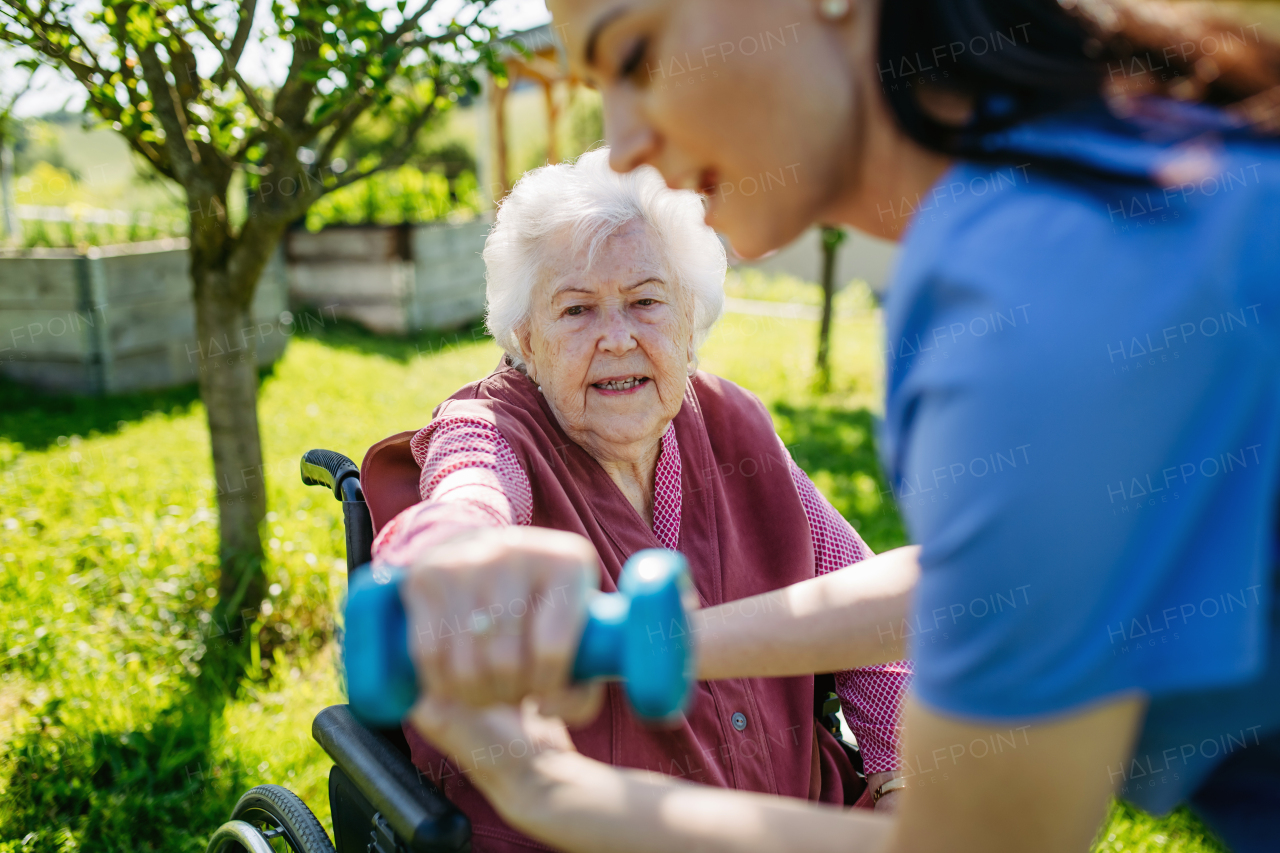 Female caregiver doing motorized exercises with senior woman in a wheelchair. Elderly woman working out with small dumbbell, nurse leading and teaching her.