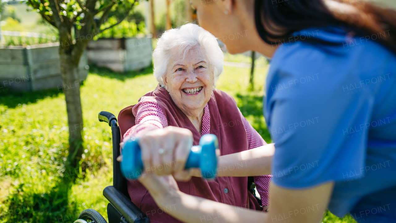 Female caregiver doing motorized exercises with senior woman in a wheelchair. Elderly woman working out with small dumbbell, nurse leading and teaching her.