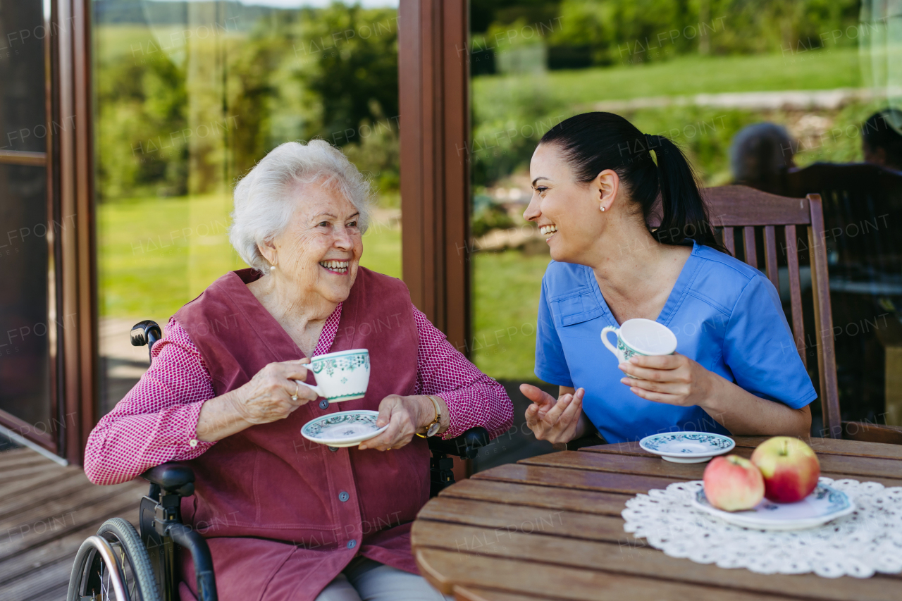 Caregiver spending time with elderly woman, drinking coffee, talking. Nurse and senior woman in wheelchair enjoying a warm day outdoors.