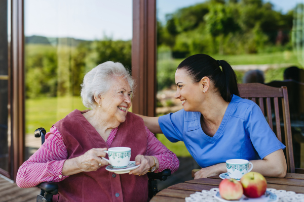 Caregiver spending time with elderly woman, drinking coffee, talking. Nurse and senior woman in wheelchair enjoying a warm day outdoors.