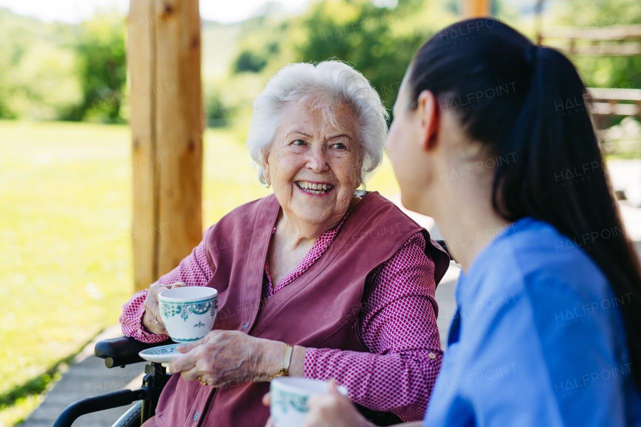 Caregiver spending time with elderly woman, drinking coffee, talking. Nurse and senior woman in wheelchair enjoying a warm day outdoors.