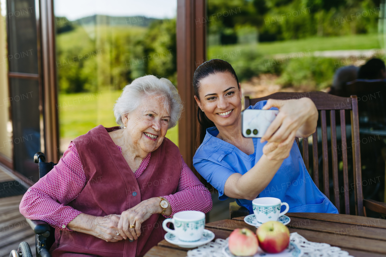 Caregiver spending time with elderly woman, drinking coffee, taking selfie. Nurse and senior woman in wheelchair enjoying a warm day outdoors.