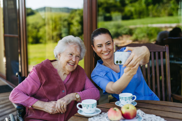 Caregiver spending time with elderly woman, drinking coffee, taking selfie. Nurse and senior woman in wheelchair enjoying a warm day outdoors.