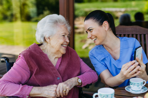 Caregiver spending time with elderly woman, drinking coffee, talking. Nurse and senior woman in wheelchair enjoying a warm day outdoors.