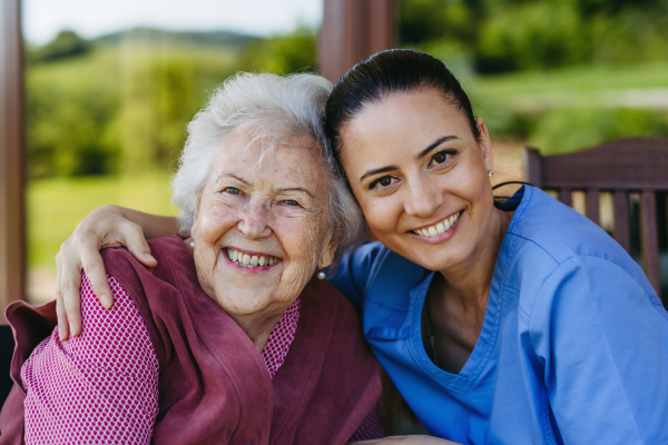 Portrait of female caregiver and senior woman embracing. Nurse and elderly woman enjoying a warm day in nursing home, public park.