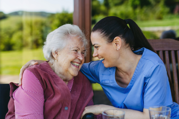 Portrait of female caregiver and senior woman embracing. Nurse and elderly woman enjoying a warm day in nursing home, public park.