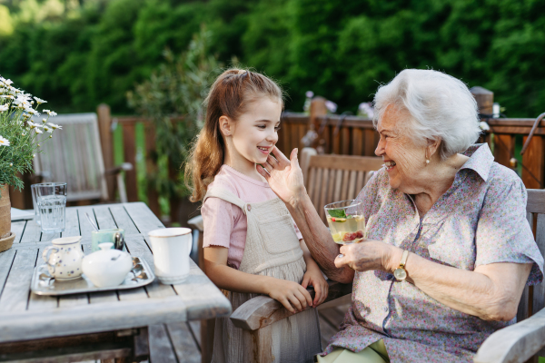 Granddaughter sitting on patio with elderly grandma, enjoying sweet iced tea. Senior lady spending time with young girl, enjoying together time.