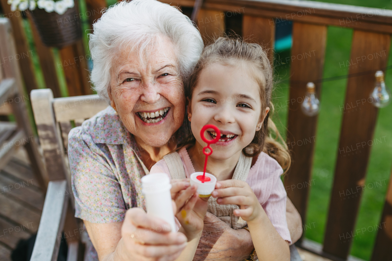 Granddaughter blowing bubbles with elderly grandma, sitting on her knees. Senior lady spending time with young girl, enjoying together time.