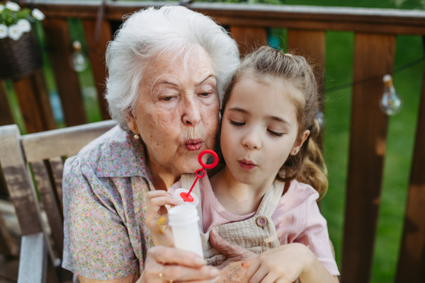 Granddaughter blowing bubbles with elderly grandma, sitting on her knees. Senior lady spending time with young girl, enjoying together time.