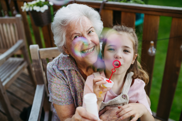 Granddaughter blowing bubbles with elderly grandma, sitting on her knees. Senior lady spending time with young girl, enjoying together time.