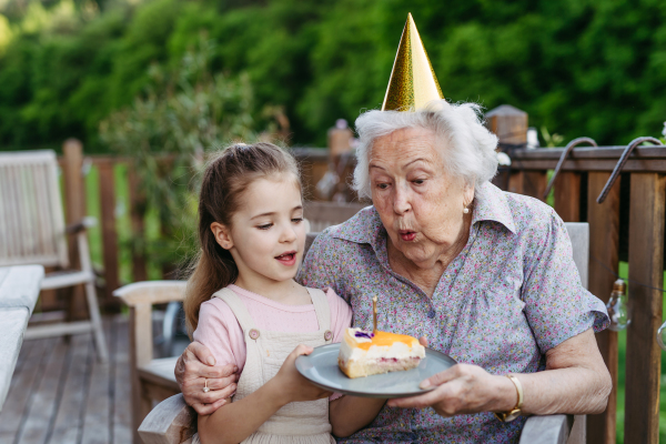 Granddaughter celebrating birthday with elderly grandma, blowing candle on cake. Weekend in cottage.