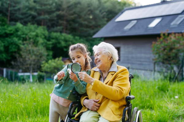 Granddaughter spending time with elderly grandma, looking at wildflowers with magnifying glass. Senior lady in a wheelchair spending time in nature.