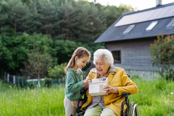 Granddaughter showing grandma model of house with solar panels on roof. Senior lady in wheelchair spending time in nature. Weekend in cottage.