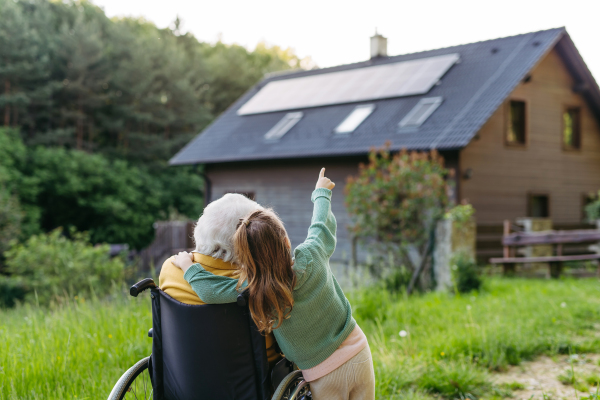 Granddaughter showing grandma solar panels on roof. Senior lady in wheelchair spending time in nature. Weekend in cottage.