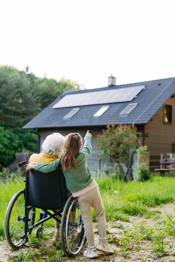 Granddaughter showing grandma solar panels on roof. Senior lady in wheelchair spending time in nature. Weekend in cottage.