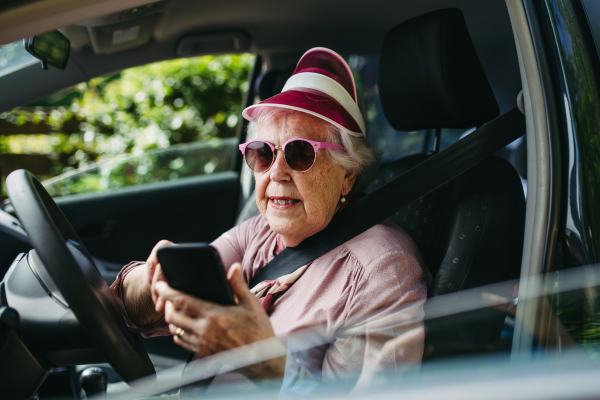 Happy senior woman driving car alone, holding smartphone, scrolling Safe driving for elderly adults, older driver safety. Driving license renewal at older age.