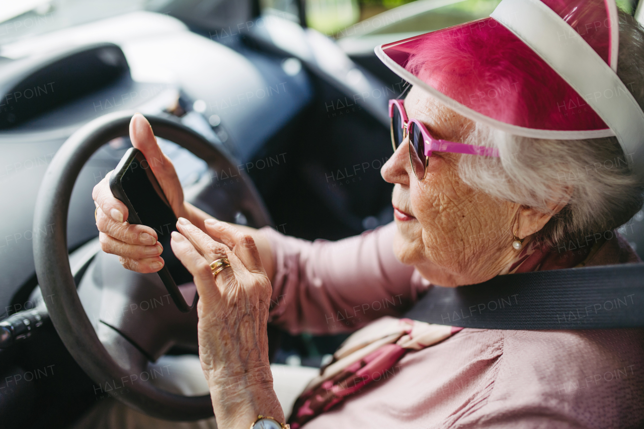 Happy senior woman driving car alone, holding smartphone, scrolling Safe driving for elderly adults, older driver safety. Driving license renewal at older age.