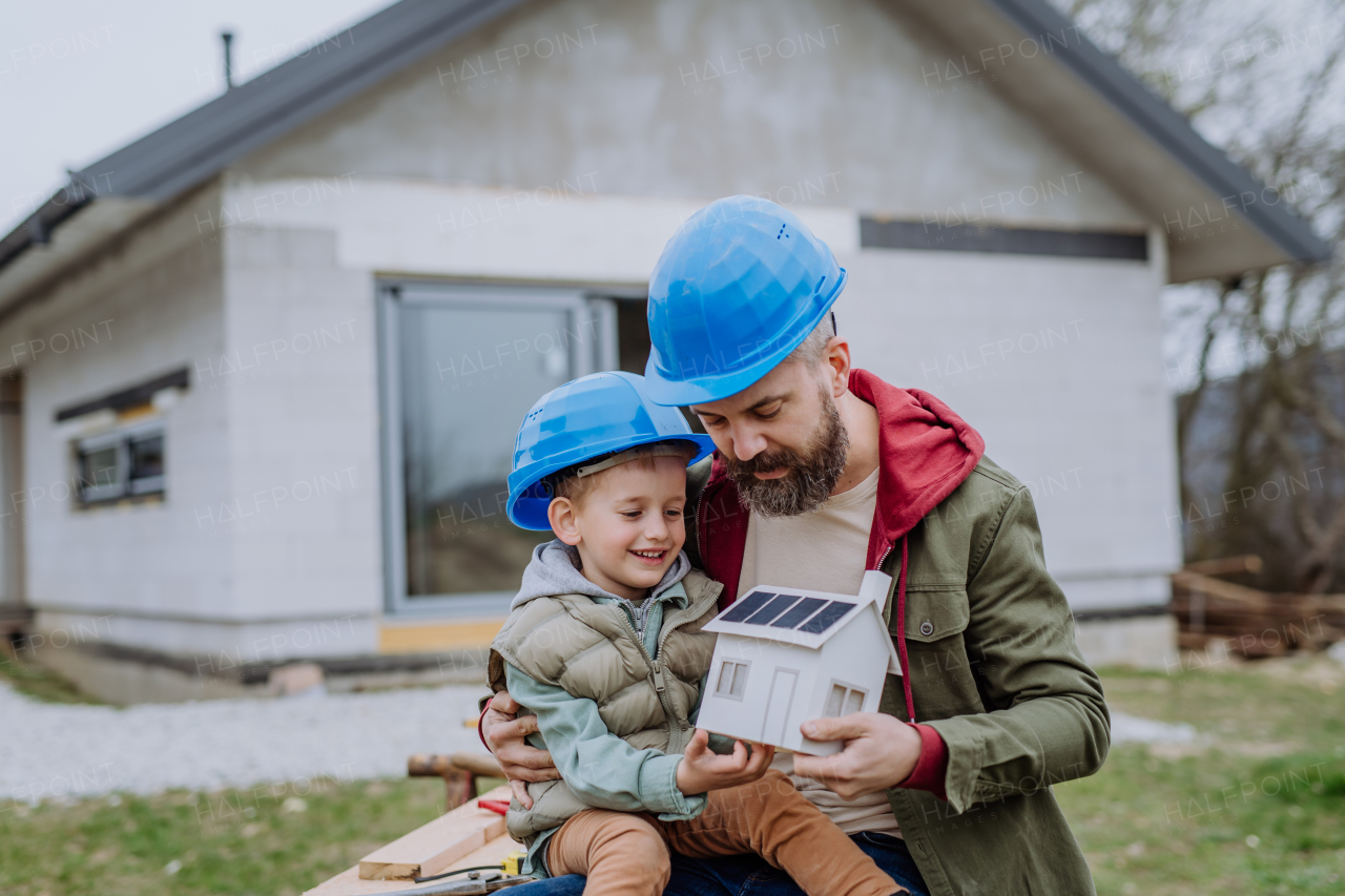 Father and his little son in front of their unfinished house holding a model of paper house with solar panels.