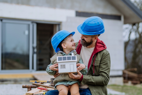 Father and his little son in front of their unfinished house holding a model of paper house with solar panels.