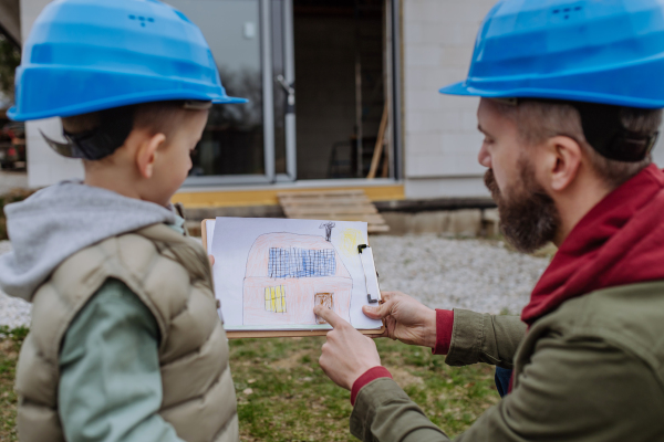 Father and his little son in front of their unfinished house holding a picture with paper house with solar panels.