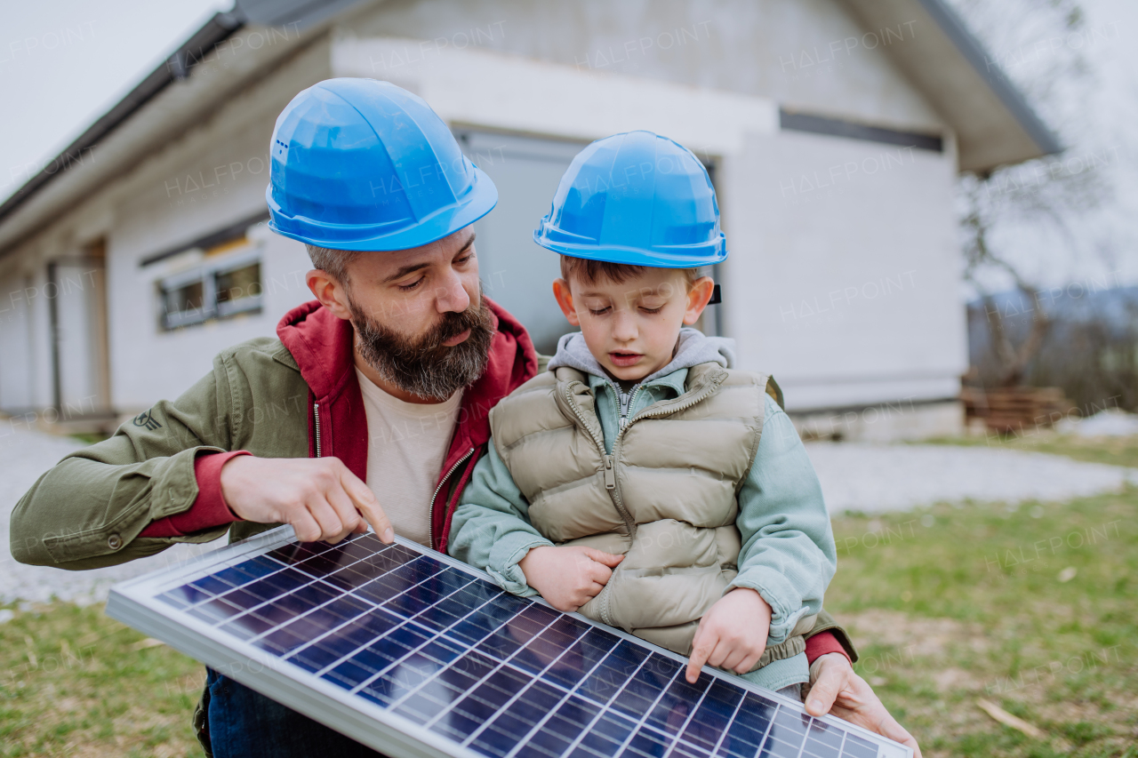 Close-up of father and his little son holding solar panel, in front of their unfinished house.