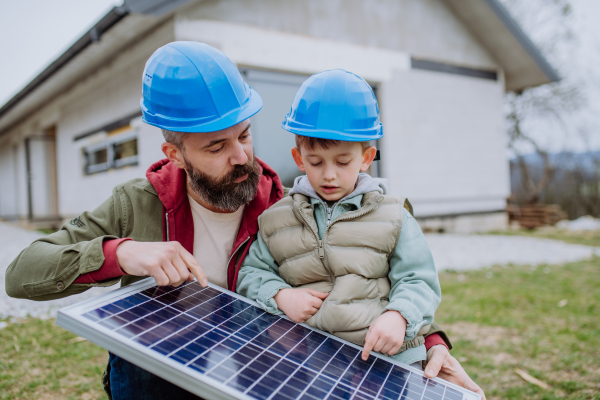 Close-up of father and his little son holding solar panel, in front of their unfinished house.