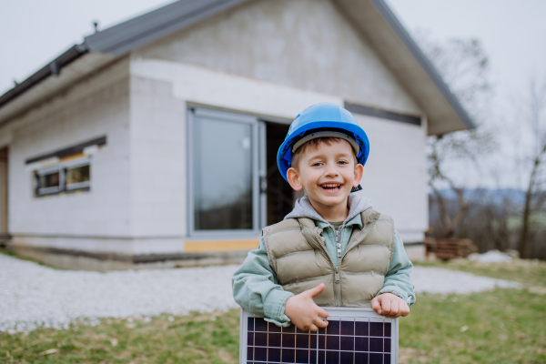 Portrait of a little boy holding solar panel, in front of their new unfinished house.