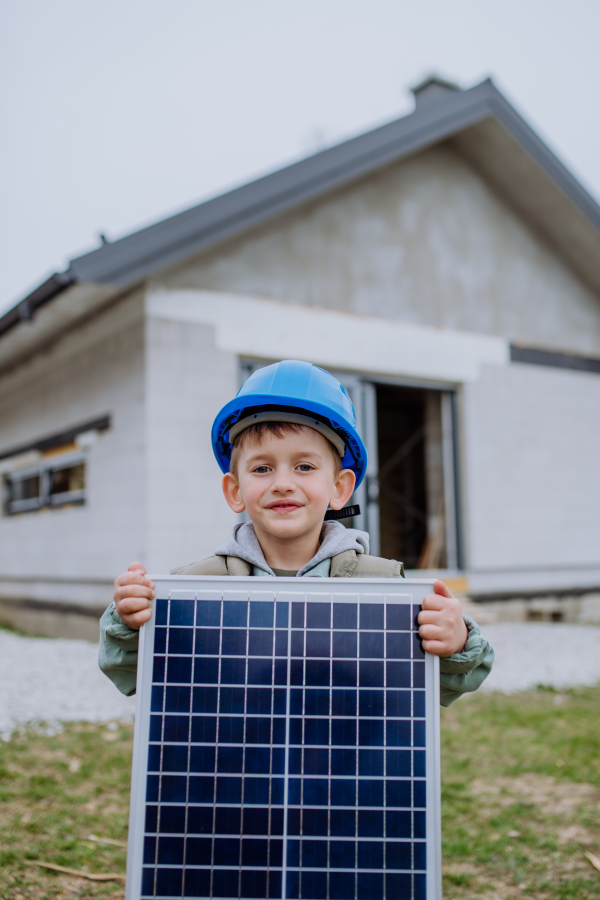 Portrait of a little boy holding solar panel, in front of their new unfinished house.