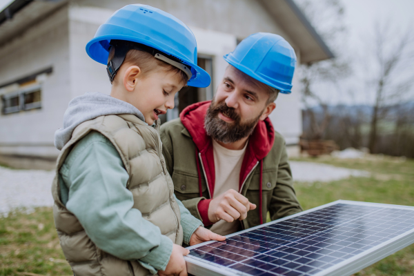 Father and his little son holding solar panel, in front of their unfinished house.