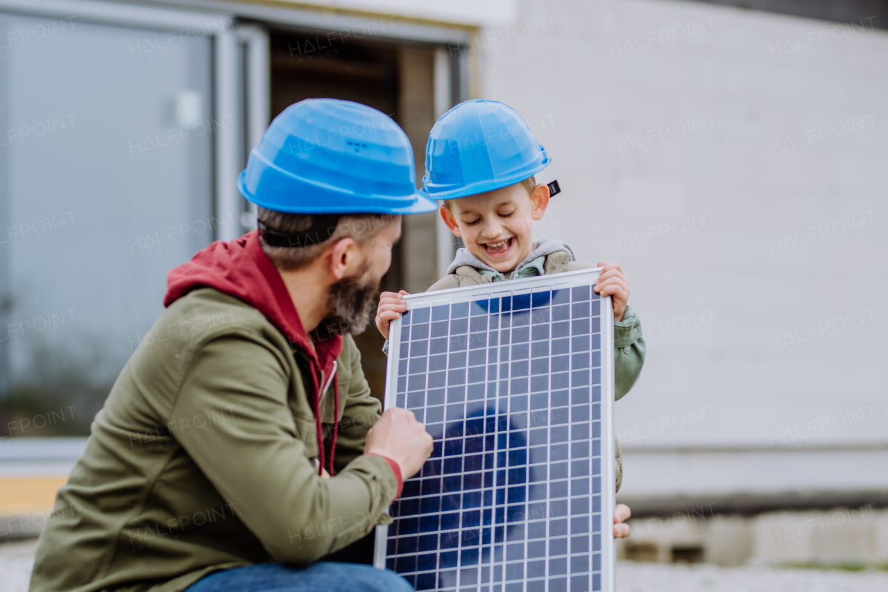 Close-up of father and his little son holding solar panel, in front of their unfinished house.