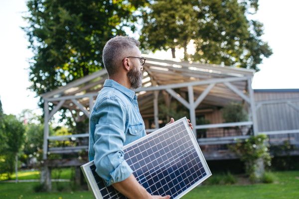 Man carrying solar panel in arms, walking across garden. Sustainable lifestyle and green household, home.