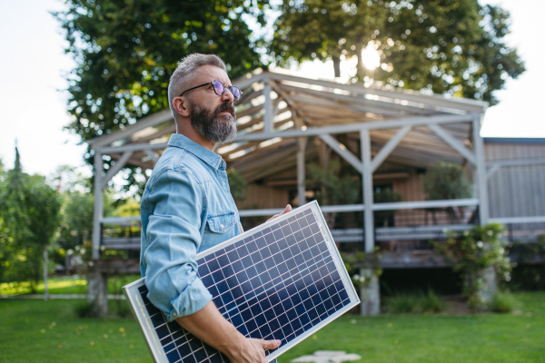 Man looking at solar panel in arms, standing in garden. Sustainable lifestyle and green household, home.