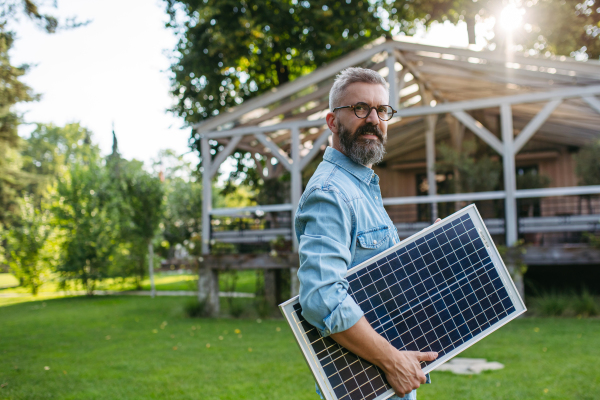 Man carrying solar panel in arms, walking across garden. Sustainable lifestyle and green household, home.