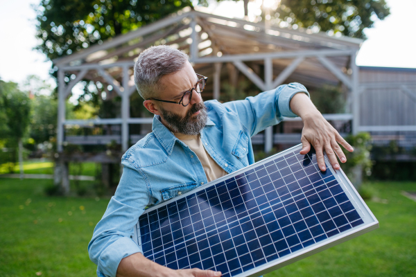 Man looking at solar panel in arms, standing in garden. Sustainable lifestyle and green household, home.