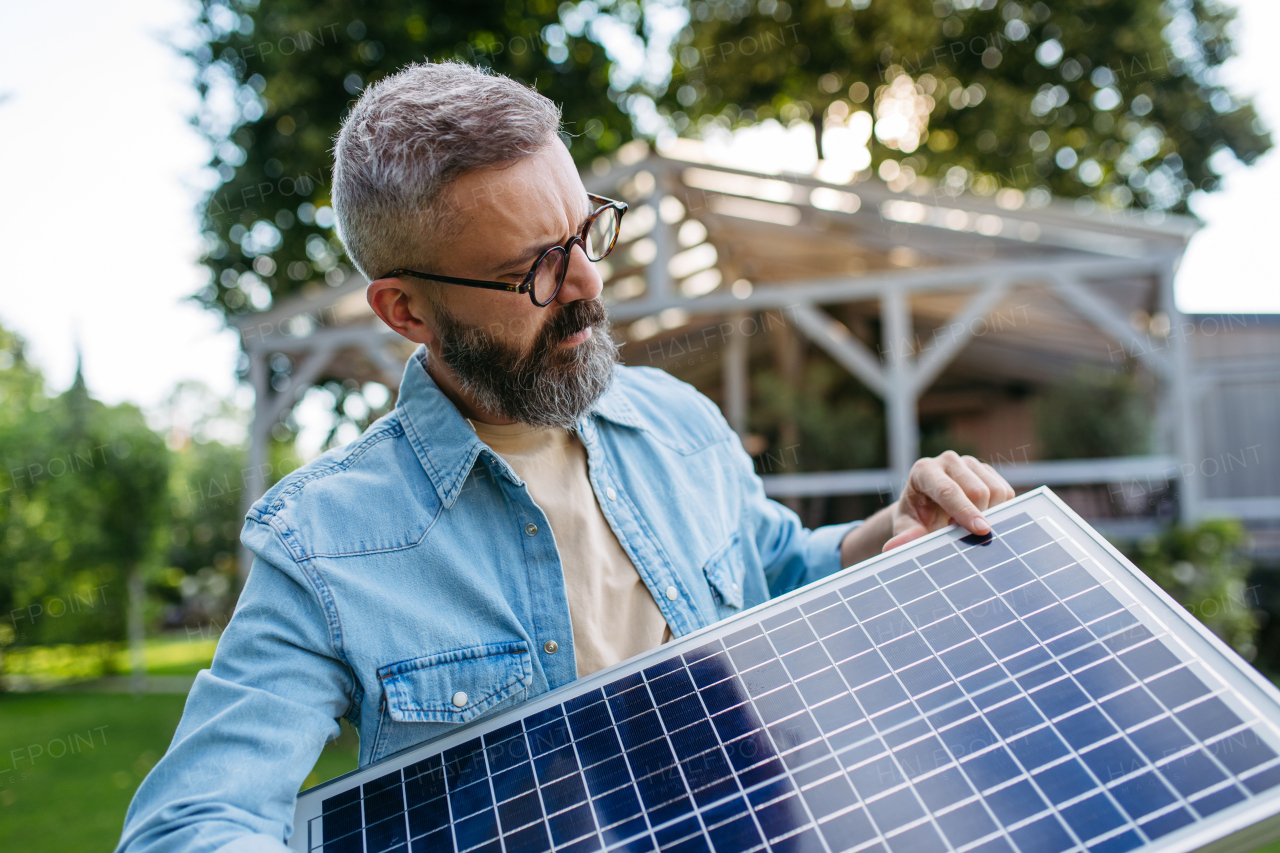 Man looking at solar panel in arms, standing in garden. Sustainable lifestyle and green household, home.