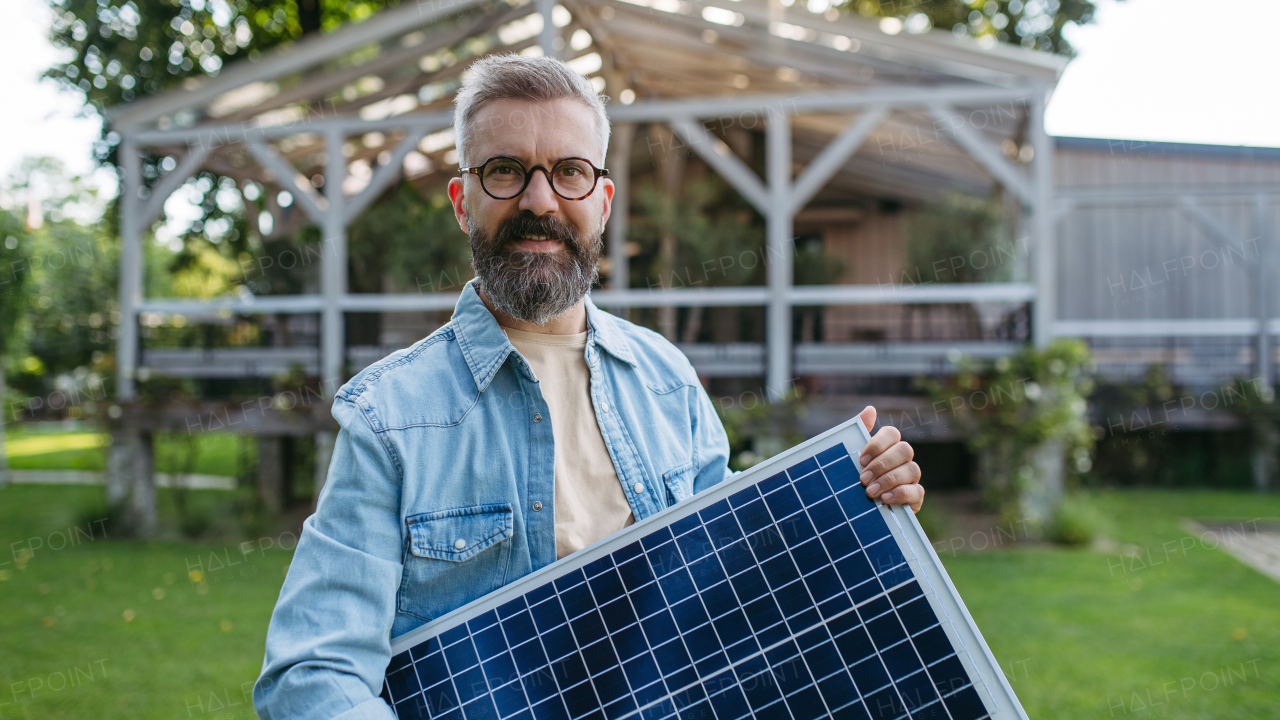 Man looking at solar panel in arms, standing in garden. Sustainable lifestyle and green household, home.