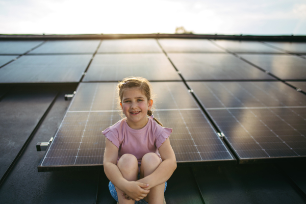 Cute girl on roof with solar panels, sitting and looking at camera. Rooftop solar or photovoltaic system. Sustainable future for next generation concept.