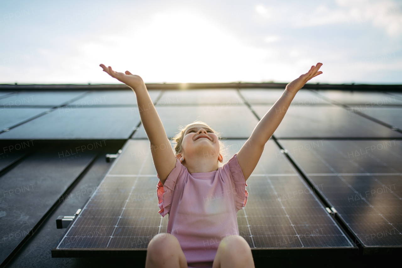 Cute girl on roof with solar panels, standing with open arms, hands up in the air. Rooftop solar or photovoltaic system. Sustainable future for next generation concept.