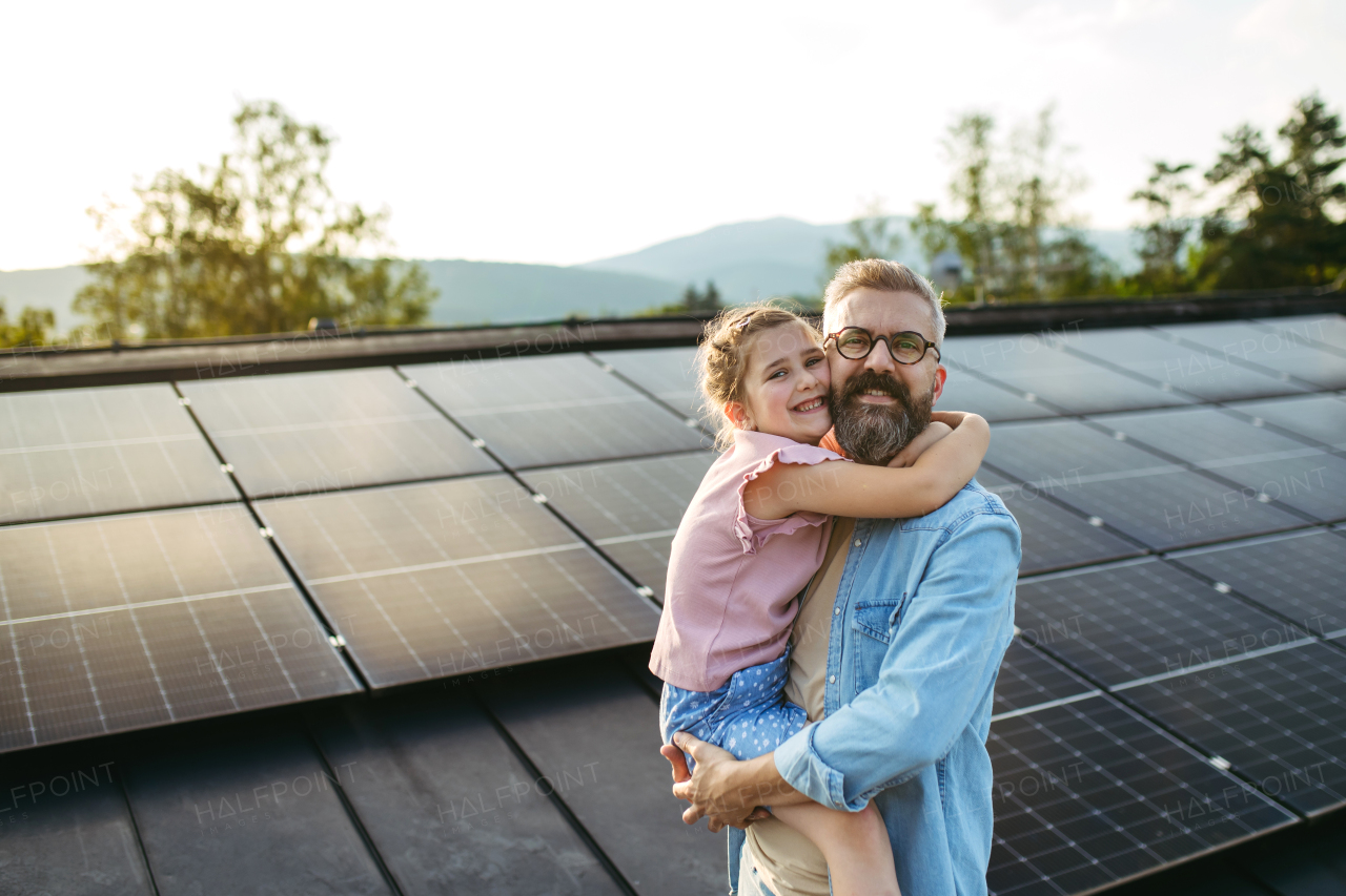 Father with girl on roof with solar panels, hugging, looking at camera. Rooftop solar or photovoltaic system. Sustainable future for next generation.