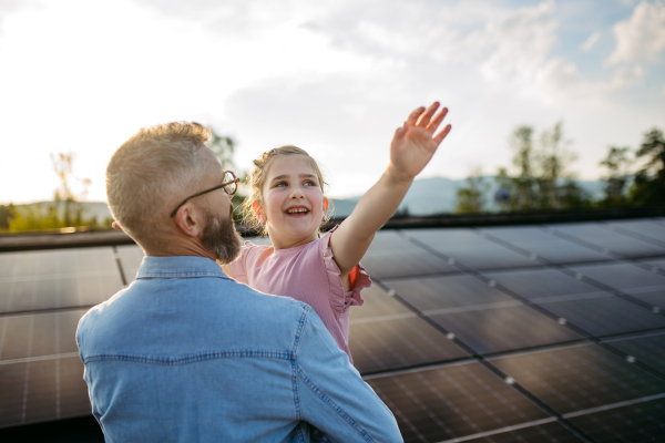 Father with girl on roof with solar panels, hugging, looking at sky. Rooftop solar or photovoltaic system. Sustainable future for next generation concept.