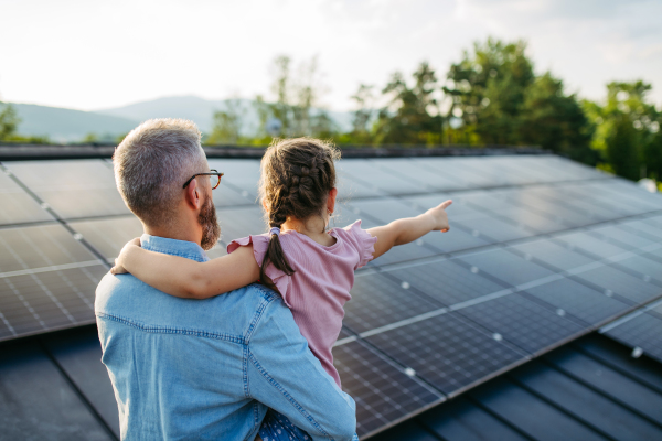 Rear view of father with girl on roof with solar panels, hugging. Rooftop solar or photovoltaic system. Sustainable future for next generation concept.